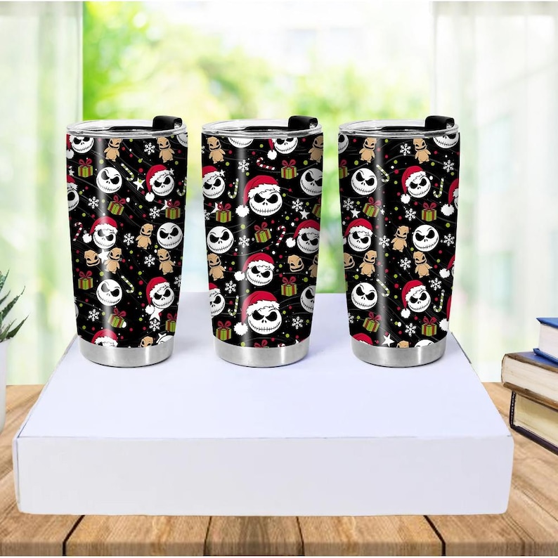 May include: Three stainless steel tumblers with black backgrounds and a repeating pattern of Jack Skellington faces wearing Santa hats, gingerbread men, presents, and snowflakes. The tumblers have black lids and silver bases.