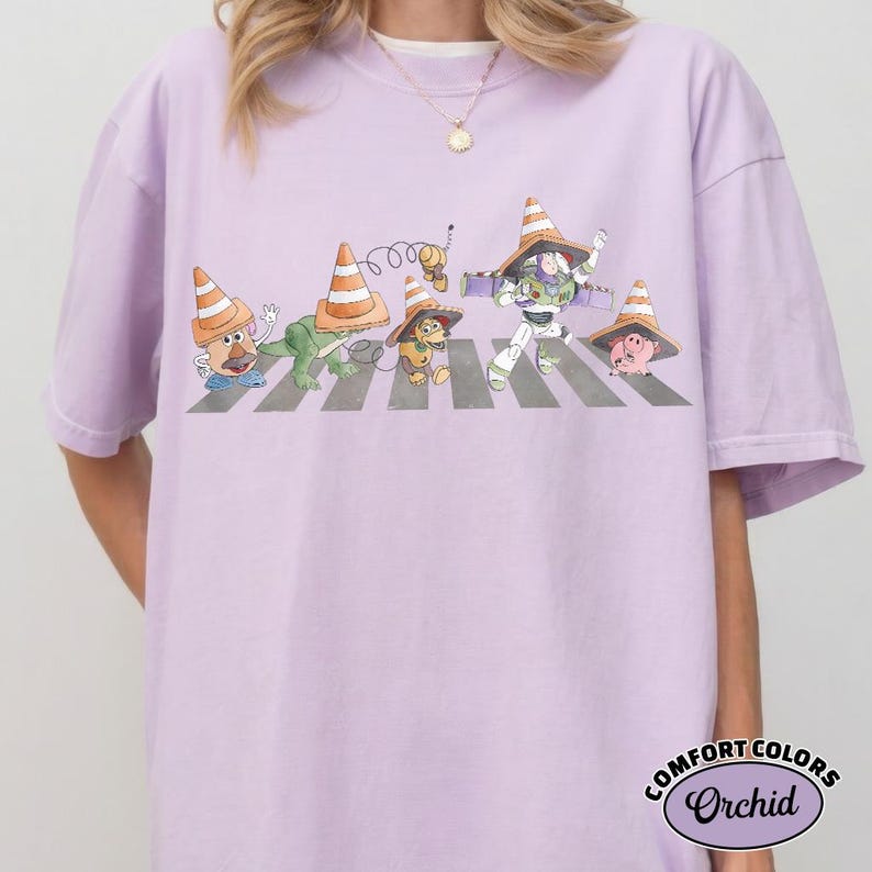 K&ouml;nnte beinhalten: Ein helllila T-Shirt mit einer Illustration von Toy Story-Charakteren, die einen Zebrastreifen &uuml;berqueren, jeder mit einem Verkehrskegel-Hut. Das Shirt hat den Text "COMFORT COLORS Orchid" in einem kreisf&ouml;rmigen Design.