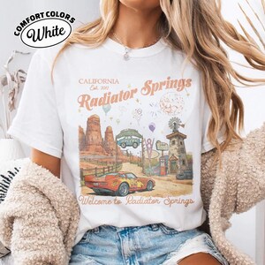Puede incluir: Camiseta blanca Comfort Colors con un gr&aacute;fico de Radiator Springs, California. El dise&ntilde;o incluye un coche vintage, un paisaje des&eacute;rtico y el texto "Welcome to Radiator Springs". La camiseta tambi&eacute;n tiene el texto "Comfort Colors White".