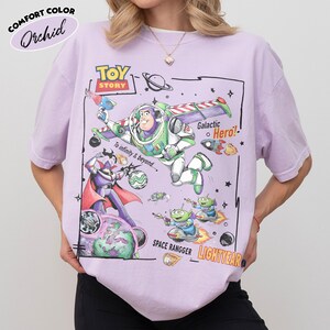 May include: A light purple t-shirt featuring a graphic print of Buzz Lightyear, Zurg, and aliens from Toy Story. The design includes the text TOY STORY, To infinity & beyond, Galactic Hero, SPACE RANGGER, and LIGHTYEAR with space-themed illustrations.