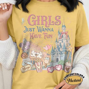 May include: Mustard-colored t-shirt with the text "GIRLS Just Wanna Have Fun" and various Disney-themed graphics. The design includes a castle, Mickey Mouse ears, popcorn, and heart-shaped sunglasses. The shirt is labeled "Comfort Colors".