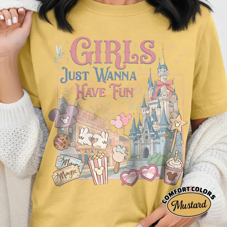 May include: Mustard-colored t-shirt with the text "GIRLS Just Wanna Have Fun" and various Disney-themed graphics. The design includes a castle, Mickey Mouse ears, popcorn, and heart-shaped sunglasses. The shirt is labeled "Comfort Colors".