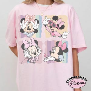 May include: Pink t-shirt featuring four panels with Minnie Mouse illustrations. Each panel shows Minnie in different poses, wearing a bow and various outfits. The shirt has a Comfort Colors Blossom label.