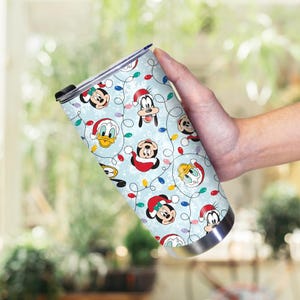 May include: A stainless steel tumbler featuring a festive pattern of Disney characters like Mickey Mouse, Donald Duck, and Goofy in Santa hats, with Christmas lights on a light blue background. The tumbler has a black lid.