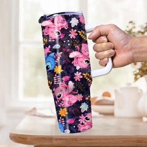 May include: A tumbler with a white handle featuring a dark background with pink and blue cartoon characters, flowers, and leaves. The tumbler is held by a hand and is on a wooden table.