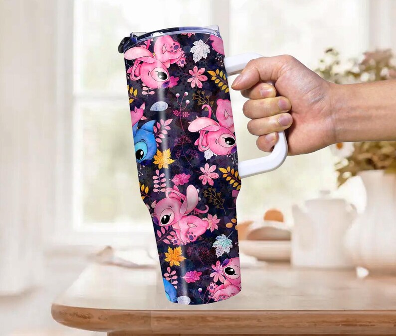 May include: A tumbler with a white handle featuring a dark background with pink and blue cartoon characters, flowers, and leaves. The tumbler is held by a hand and is on a wooden table.