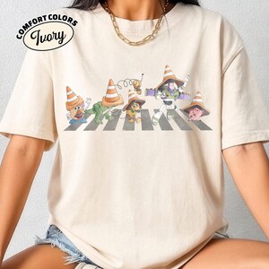K&ouml;nnte beinhalten: Elfenbeinfarbenes Comfort Colors T-Shirt mit einer Parodie des Abbey Road-Albumcovers der Beatles. Das Design zeigt Toy Story-Charaktere, die einen Zebrastreifen &uuml;berqueren, jeder mit einem Verkehrskegel-Hut. Das Shirt hat eine entspannte Passform.