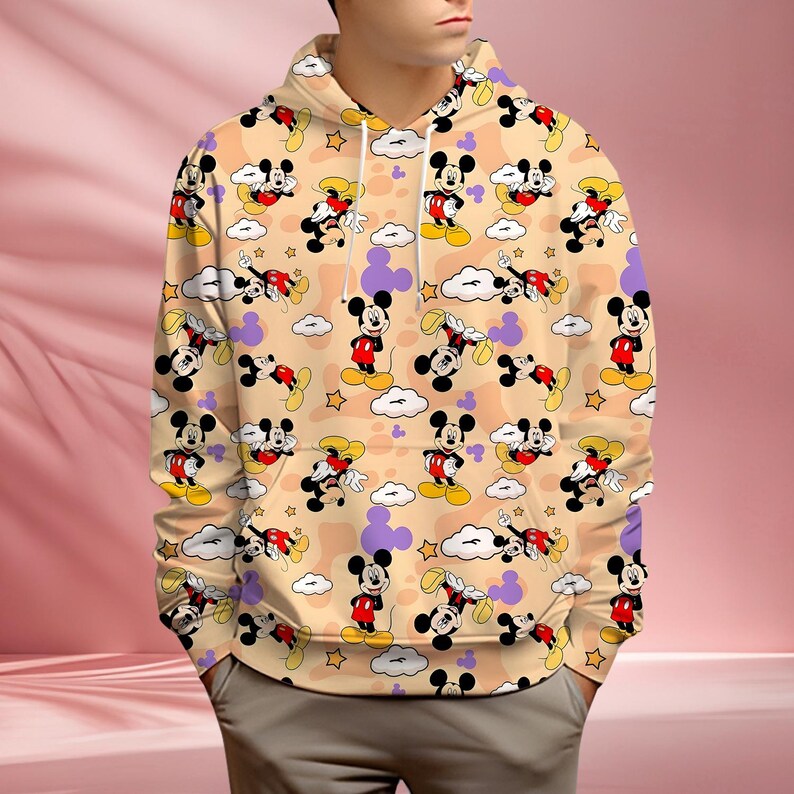K&ouml;nnte beinhalten: Beiger Kapuzenpullover mit sich wiederholendem Muster von Mickey Mouse, Wolken und Sternen. Mickey ist in seinen klassischen roten Shorts, gelben Schuhen und schwarzen Handschuhen dargestellt. Der Hintergrund hat eine pfirsichfarbene Farbe.