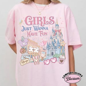 May include: A light pink t-shirt with the text "GIRLS JUST WANNA HAVE FUN" and a graphic of a castle, fireworks, and other theme park icons. The shirt also features the Comfort Colors Blossom logo.