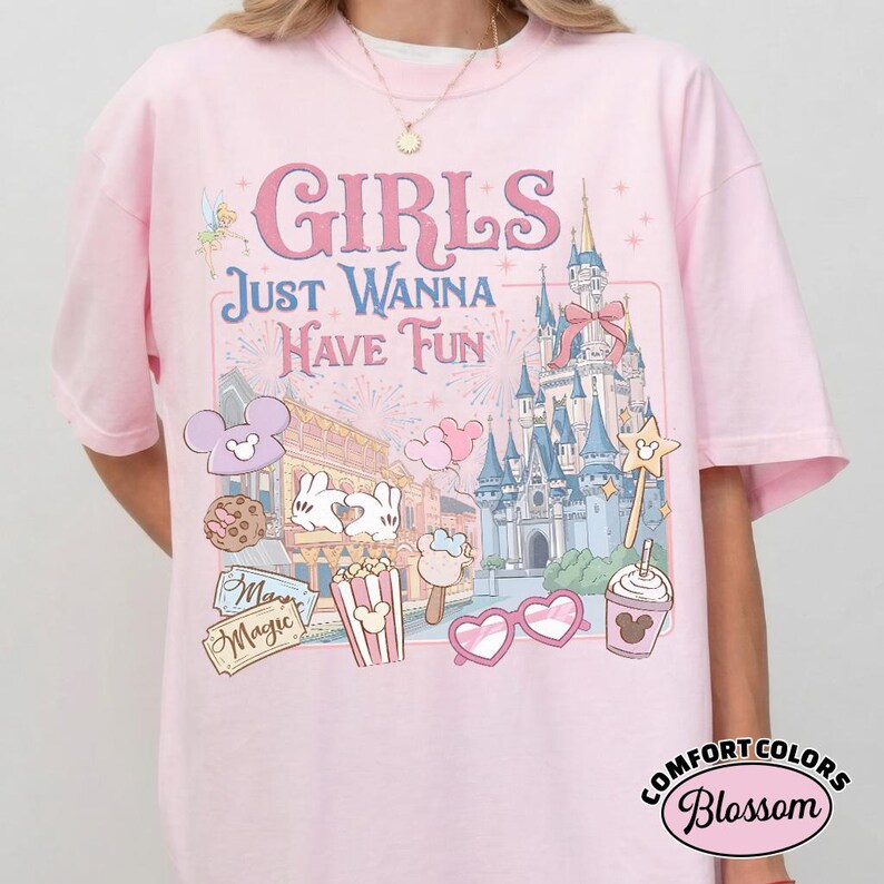 May include: A light pink t-shirt with the text "GIRLS JUST WANNA HAVE FUN" and a graphic of a castle, fireworks, and other theme park icons. The shirt also features the Comfort Colors Blossom logo.