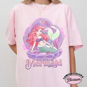 Op de afbeelding: Lichtroze T-shirt met een cartoon illustratie van een zeemeermin met roze haar, een paarse schelp en de tekst "The Little Mermaid". De zeemeermin draagt een paarse top en heeft een groene staart. Het shirt heeft een ronde hals.