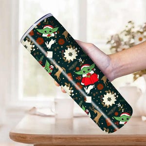 Pode incluir: Um copo de a&ccedil;o inoxid&aacute;vel com um design festivo de Natal. O copo apresenta um fundo verde escuro com imagens repetidas de um personagem alien&iacute;gena verde usando um chap&eacute;u de Papai Noel e um casaco vermelho, renas e estrelas brancas. O copo &eacute; segurado por uma pessoa.
