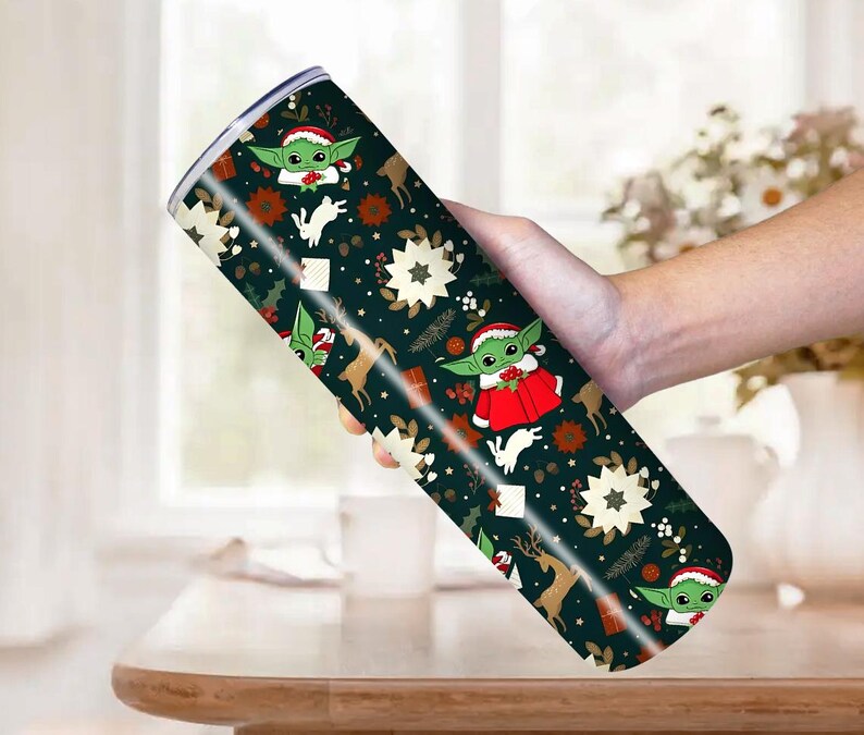 Pode incluir: Um copo de a&ccedil;o inoxid&aacute;vel com um design festivo de Natal. O copo apresenta um fundo verde escuro com imagens repetidas de um personagem alien&iacute;gena verde usando um chap&eacute;u de Papai Noel e um casaco vermelho, renas e estrelas brancas. O copo &eacute; segurado por uma pessoa.