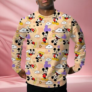 K&ouml;nnte beinhalten: Beiges Sweatshirt mit einem sich wiederholenden Muster von Mickey Mouse-Charakteren in verschiedenen Posen. Das Design umfasst wei&szlig;e Wolken, gelbe Sterne und lila Formen. Das lang&auml;rmlige Oberteil ist ein l&auml;ssiger Stil.