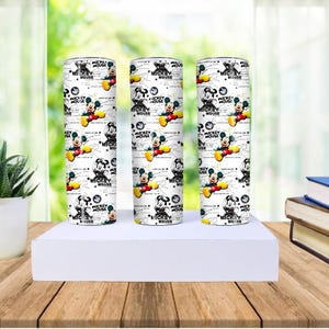 May include: Three white tumblers with a repeating pattern of Mickey Mouse in various poses. The design includes the text "MICKEY MOUSE" and other graphics. The tumblers are cylindrical and stand on a white surface.
