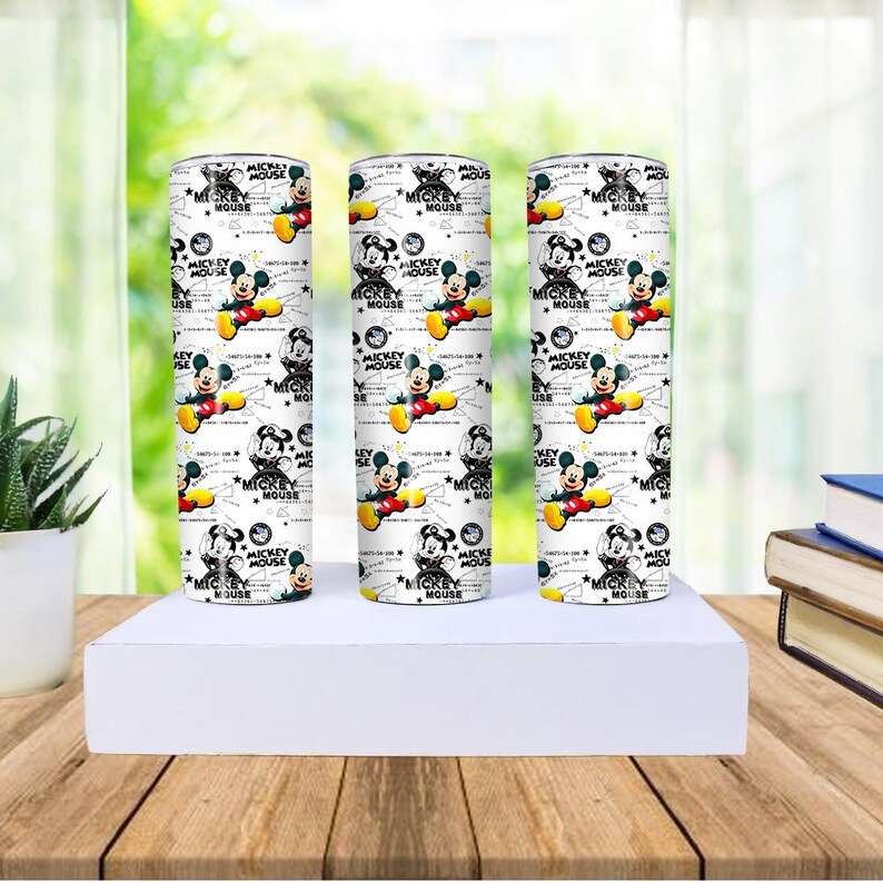 May include: Three white tumblers with a repeating pattern of Mickey Mouse in various poses. The design includes the text "MICKEY MOUSE" and other graphics. The tumblers are cylindrical and stand on a white surface.