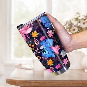 May include: A stainless steel tumbler featuring a colorful design with the character Stitch from Lilo & Stitch. The design includes blue Stitch characters, pink flowers, and autumn leaves on a dark background. The tumbler is held by a person.
