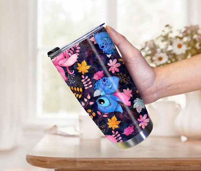 May include: A stainless steel tumbler featuring a colorful design with the character Stitch from Lilo & Stitch. The design includes blue Stitch characters, pink flowers, and autumn leaves on a dark background. The tumbler is held by a person.