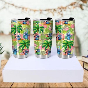 May include: Three stainless steel travel tumblers with black lids. Each tumbler features a colorful tropical design with palm trees, flowers, and cartoon characters. The tumblers are approximately 7 inches tall.