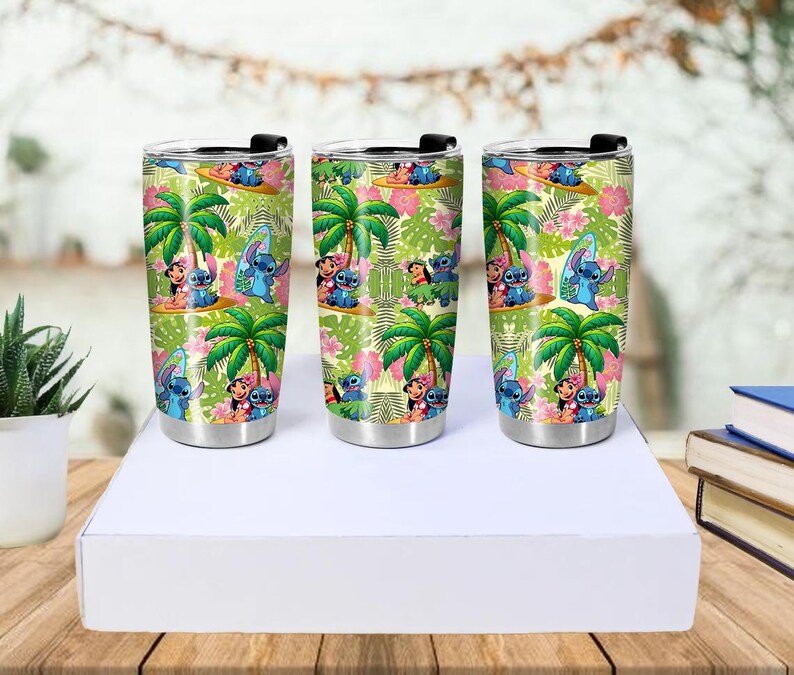 May include: Three stainless steel travel tumblers with black lids. Each tumbler features a colorful tropical design with palm trees, flowers, and cartoon characters. The tumblers are approximately 7 inches tall.