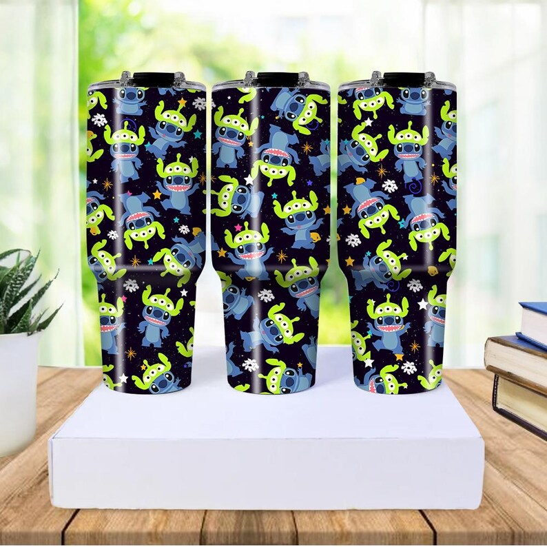 Peut inclure: Trois gobelets en acier inoxydable avec des couvercles noirs et un motif enveloppant avec des personnages de dessins anim&eacute;s bleus et verts sur un fond bleu fonc&eacute;. Les gobelets sont cylindriques et reposent sur une surface blanche.
