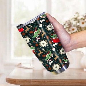 Pode incluir: Um copo de a&ccedil;o inoxid&aacute;vel com um design de Natal festivo. O copo apresenta um fundo verde escuro com padr&otilde;es repetidos de um personagem alien&iacute;gena verde, renas, flocos de neve e presentes. O personagem est&aacute; usando um chap&eacute;u de Papai Noel e roupas vermelhas.