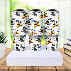 May include: Three white insulated tumblers with a repeating pattern of Mickey Mouse illustrations. The design includes Mickey Mouse in various poses, along with the words "MICKEY MOUSE" and stars. The tumblers are on a white surface.