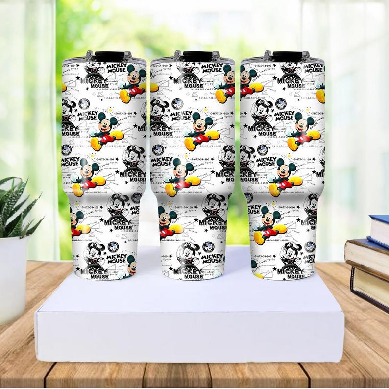 May include: Three white insulated tumblers with a repeating pattern of Mickey Mouse illustrations. The design includes Mickey Mouse in various poses, along with the words "MICKEY MOUSE" and stars. The tumblers are on a white surface.