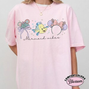 May include: Light pink t-shirt with a graphic design featuring a cartoon fish, seashell headband, and a seashell-shaped ice cream cone. The words "Mermaid vibes" are written in cursive. The shirt has a Comfort Colors Blossom label.
