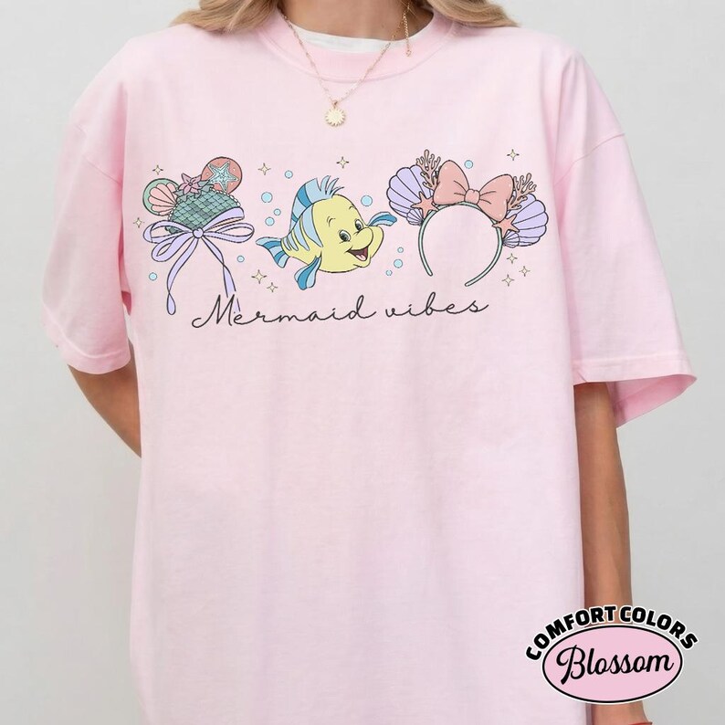May include: Light pink t-shirt with a graphic design featuring a cartoon fish, seashell headband, and a seashell-shaped ice cream cone. The words "Mermaid vibes" are written in cursive. The shirt has a Comfort Colors Blossom label.