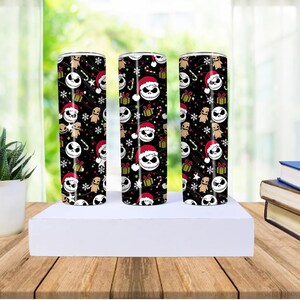 May include: Three black insulated tumblers with a repeating pattern of Jack Skellington faces wearing Santa hats, gingerbread men, and candy canes. The design includes green gift boxes and white stars on a black background. The tumblers are cylindrical.