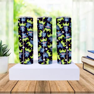 Peut inclure: Trois gobelets cylindriques avec un fond noir, pr&eacute;sentant un motif r&eacute;p&eacute;t&eacute; de personnages de dessins anim&eacute;s bleus et verts. Les gobelets sont expos&eacute;s sur une surface blanche, avec une table en bois et une plante en arri&egrave;re-plan.
