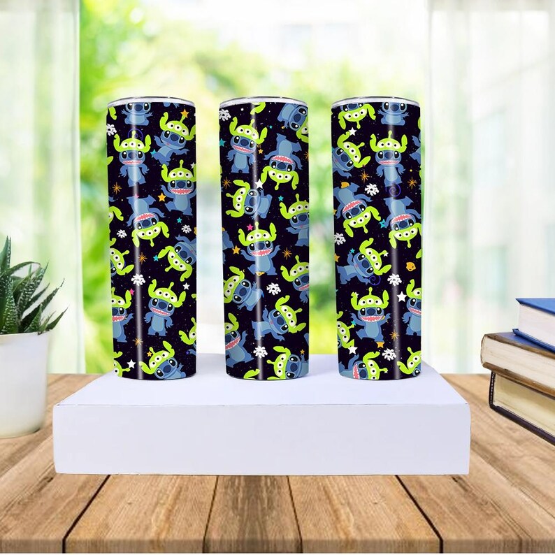 Peut inclure: Trois gobelets cylindriques avec un fond noir, pr&eacute;sentant un motif r&eacute;p&eacute;t&eacute; de personnages de dessins anim&eacute;s bleus et verts. Les gobelets sont expos&eacute;s sur une surface blanche, avec une table en bois et une plante en arri&egrave;re-plan.