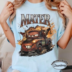 May include: Pale blue t-shirt with a graphic of Mater from the Cars film, with the name "MATER" above the image. The design includes a brown tow truck with a smiling face and lightning bolt accents. The shirt is a Comfort Colors Chambray.