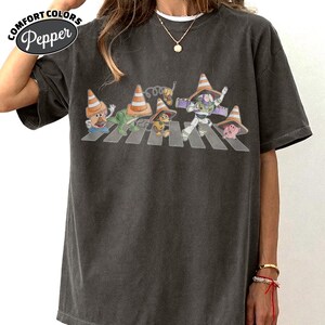 K&ouml;nnte beinhalten: Dunkelgraues Comfort Colors T-Shirt mit einer Parodie des Abbey Road-Albumcovers. Das Design zeigt Zeichentrickfiguren, die einen Zebrastreifen &uuml;berqueren, jede mit einem Verkehrskegel-Hut. Das Shirt hat die Comfort Colors und Pepper Logos.