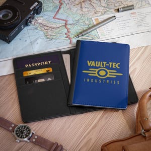 May include: Black leather passport holder with a blue cover featuring the Vault-Tec Industries logo in yellow. The open wallet displays a passport and credit cards. A watch, camera, and pen are also visible.