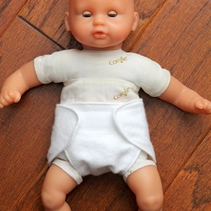 May include: A white cloth diaper with the Corolle logo on the front, worn by a baby doll with a light brown complexion and blue eyes.