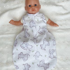 May include: A baby doll wearing a white sleep sack with a gray elephant pattern and green leaf accents. The doll has blue eyes and is lying on a white, textured surface.
