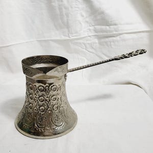 以下が含まれることがあります： 銀色のトルココーヒーポット。装飾的な花柄模様が施されています。ポットには長く装飾的なハンドルと、上部に向かって細くなる広いベースがあります。背景は白いシーツです。