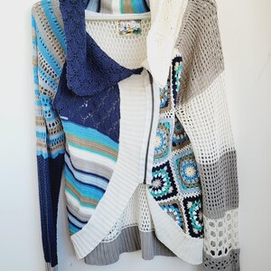 Könnte beinhalten: Eine mehrfarbige Strickjacke mit offenem Frontdesign und Reißverschluss. Der Pullover hat ein Patchwork-Design mit Blau-, Weiß-, Grau- und Brauntönen. Die Ärmel sind lang und der Kragen ist im Schal-Stil. Das Kleidungsstück hängt an einem weißen Kleiderbügel.