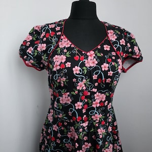 May include: Black dress with a floral pattern of pink flowers and red cherries. The dress has a V-neckline and short sleeves with red trim. The dress is displayed on a black mannequin.