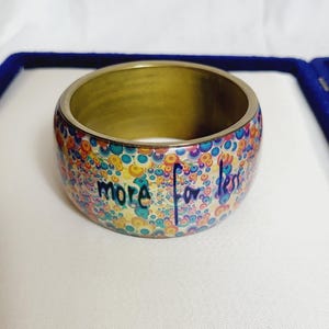May include: A wide, gold-toned bangle bracelet with a colorful, dotted pattern in shades of blue, yellow, and purple. The bracelet has the words "more for less" in a handwritten style. The bracelet is displayed on a white surface.