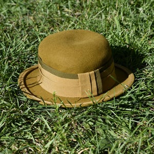 May include: A vintage-style olive green hat with a rounded crown and a wide brim. The hat features a contrasting tan band around the base and a decorative olive green ribbon. The hat is resting on a bed of green grass.