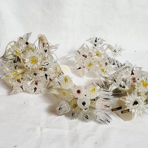 May include: Clear plastic Christmas light strands with starburst-shaped bulbs. Each bulb has a central yellow or red accent, surrounded by clear, spiky points. The lights are strung on white cords, and the plugs are visible.