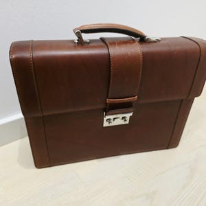 May include: A structured brown leather briefcase featuring a top handle, a vertical strap closure, and a silver-toned metal push-lock mechanism. The rectangular bag has clean lines, visible stitching along the edges, and a smooth, polished finish.