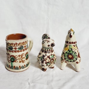 Op de afbeelding: Drie keramische beeldjes met bloemen en geometrische ontwerpen. De set bevat een mok, een konijn en een paard, allemaal beschilderd in crème, bruin, groen en rood. De mok heeft een handvat en een decoratieve rand. Het konijn en het paard staan.