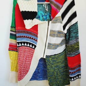 May include: A colorful patchwork zip-up sweater with a variety of patterns and textures. The sweater features a mix of red, blue, green, yellow, and white panels, with a high collar and a metal zipper. The label reads "Desigual".