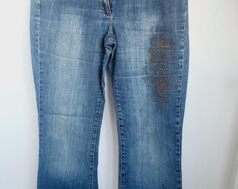 May include: Blue denim jeans with a faded wash and a slight flare at the hem. Features decorative embroidery on the right leg and red stitching details on the pockets. The jeans are hanging on a hanger.