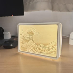 May include: A rectangular, white-framed light box featuring a relief of Hokusai's "The Great Wave off Kanagawa." The artwork is illuminated with a warm, yellow glow. The light box sits on a wooden desk.
