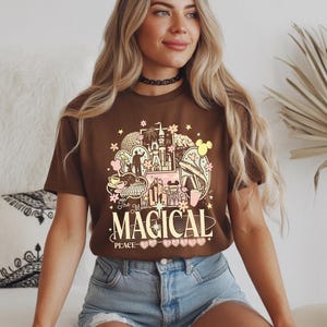 Może przedstawiać: Brązowa koszulka z kolorowym nadrukiem graficznym. Projekt zawiera ilustracje zamku, kwiat&oacute;w i innych fantazyjnych element&oacute;w z napisem "The Magical Place". Koszulka wykonana jest z miękkiego materiału.